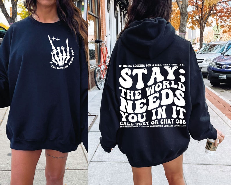 Puede incluir: Sudadera negra con texto blanco. La parte delantera presenta un gesto de mano de esqueleto y las palabras "The Worlds Needs You". La parte trasera de la sudadera con capucha dice "Stay; The World Needs You In It" con detalles de prevenci&oacute;n del suicidio.