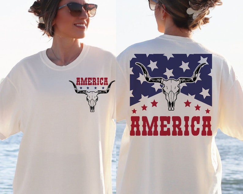 May include: White t-shirt with a red, white, and blue American flag design featuring a bull skull and the word "AMERICA" printed on the back. The front of the shirt has a smaller version of the bull skull design with the word "AMERICA" printed above it.