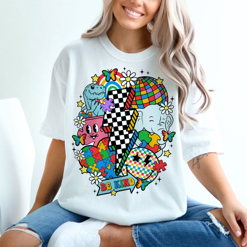 Op de afbeelding: Wit T-shirt met een kleurrijk grafisch ontwerp met een bliksem, een dinosaurus, een olifant, een puzzelhart en een discobal. De tekst "BE KIND" is onder het ontwerp gedrukt.