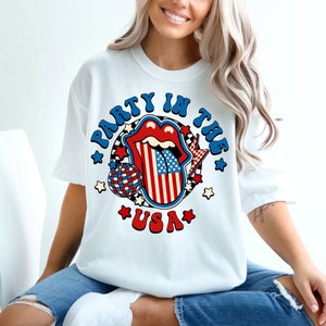 May include: White t-shirt with a graphic of a red, white, and blue tongue with a disco ball and stars. The text "Party in the USA" is printed around the tongue.