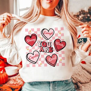 May include: White sweatshirt with a pink and white checkered background. The words "You Are" are in large, red letters. There are five pink hearts with words on them: "Strong", "Enough", "Worthy", "Capable", and "Loved".