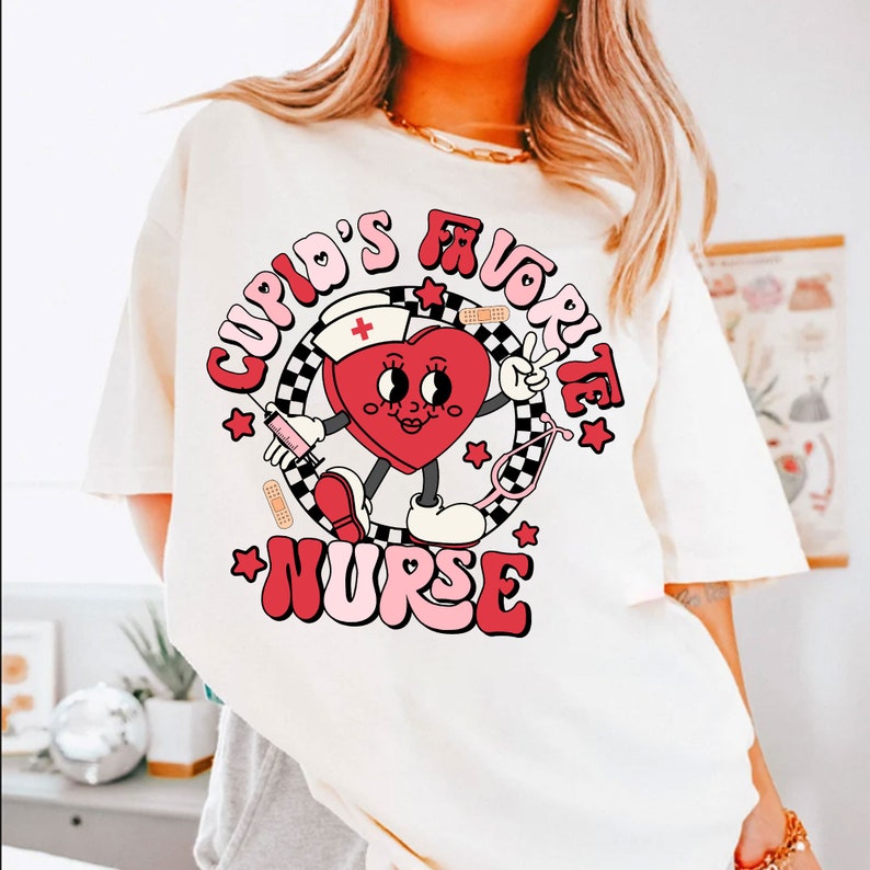 Op de afbeelding: Wit T-shirt met een rode hartgrafiek. Het hart heeft een gezicht en houdt een vredesteken vast. De tekst rond het hart zegt "Cupid's Favorite Nurse".