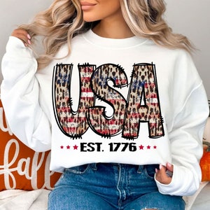 May include: White sweatshirt with a graphic of the word "USA" in red, white, and blue with a leopard print pattern. The text "EST. 1776" is below the graphic.