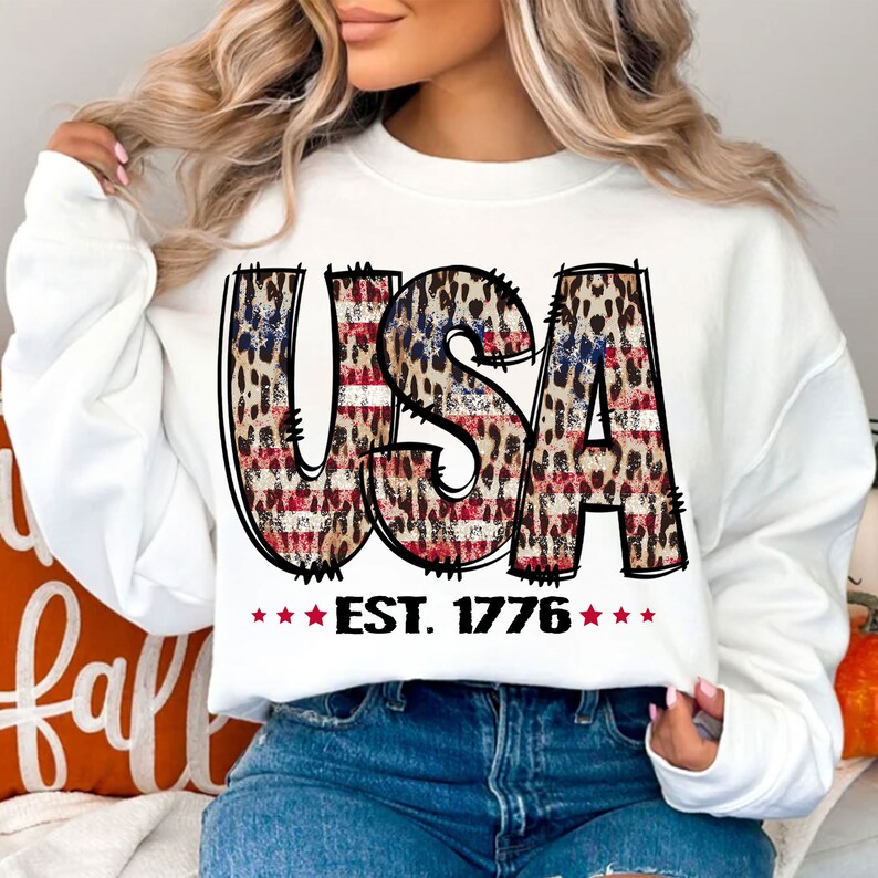 May include: White sweatshirt with a graphic of the word "USA" in red, white, and blue with a leopard print pattern. The text "EST. 1776" is below the graphic.