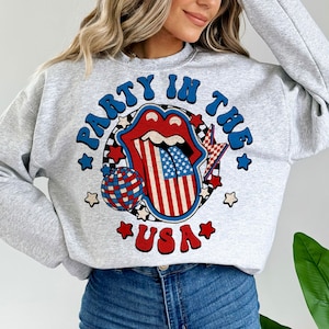 May include: Gray sweatshirt with a graphic of a red, white, and blue tongue with a disco ball and stars. The text "Party in the USA" is printed on the sweatshirt.