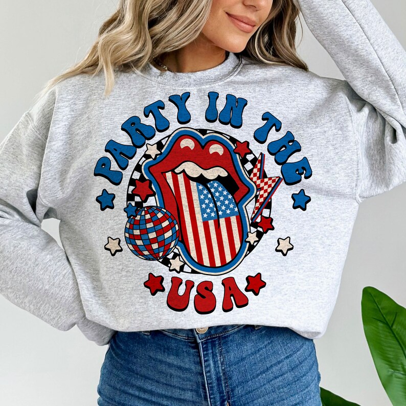 May include: Gray sweatshirt with a graphic of a red, white, and blue tongue with a disco ball and stars. The text "Party in the USA" is printed on the sweatshirt.