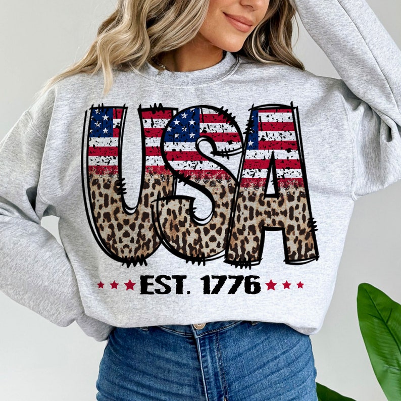 May include: Gray sweatshirt with a graphic of the word "USA" in red, white, and blue with a leopard print design. The text "EST. 1776" is below the graphic.