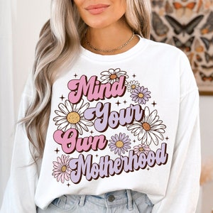 May include: White long-sleeve sweatshirt with a pink and purple graphic design that says "Mind Your Own Motherhood" surrounded by white daisies with pink and purple centers.