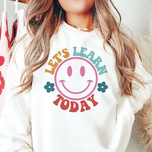 以下が含まれることがあります： スマイリーフェイスと「Let's Learn Today」のテキストが描かれたカラフルなグラフィックデザインの白いスウェットシャツ。