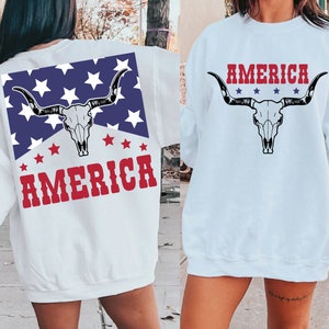 May include: White sweatshirt with a red, white, and blue American flag design on the back. The design features a cow skull with horns and the word "AMERICA" in red letters. The front of the sweatshirt has a cow skull with horns and the word "AMERICA" in red letters.