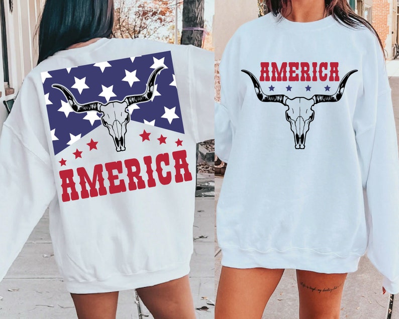 May include: White sweatshirt with a red, white, and blue American flag design on the back. The design features a cow skull with horns and the word "AMERICA" in red letters. The front of the sweatshirt has a cow skull with horns and the word "AMERICA" in red letters.