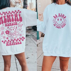 May include: Two white sweatshirts with pink graphic designs. The first sweatshirt has the text "Mental Health Matters" with a checkered pattern and smiley faces. The second sweatshirt has the text "Be Kind to Your Mind" with a smiley face.