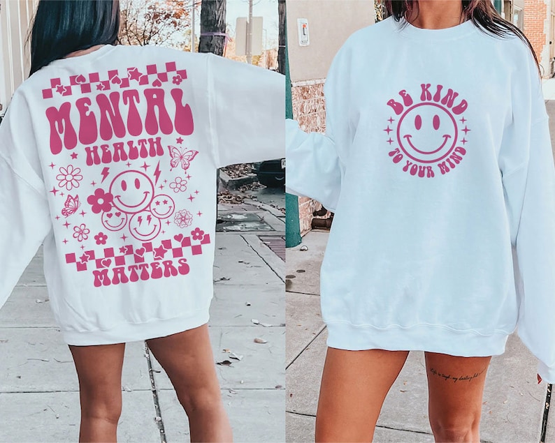 May include: Two white sweatshirts with pink graphic designs. The first sweatshirt has the text "Mental Health Matters" with a checkered pattern and smiley faces. The second sweatshirt has the text "Be Kind to Your Mind" with a smiley face.