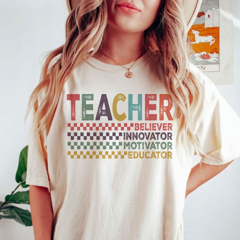 Peut inclure: Un t-shirt blanc avec le mot "TEACHER" en lettres r&eacute;tro color&eacute;es. Sous le mot se trouvent les mots "BELIEVER", "INNOVATOR", "MOTIVATOR" et "EDUCATOR" dans un motif &agrave; carreaux.