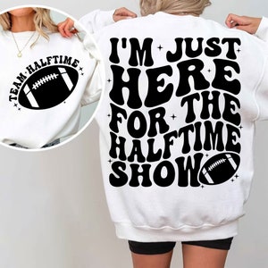 May include: White sweatshirt with black text that reads "I'm Just Here For The Halftime Show" in a retro font. A black football graphic is below the text.