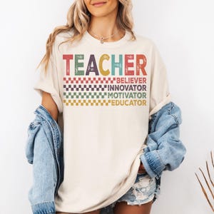 Peut inclure: Un t-shirt beige avec le mot "TEACHER" en lettres color&eacute;es et les mots "BELIEVER", "INNOVATOR", "MOTIVATOR" et "EDUCATOR" empil&eacute;s en dessous. Le t-shirt a un motif &agrave; carreaux derri&egrave;re les mots.