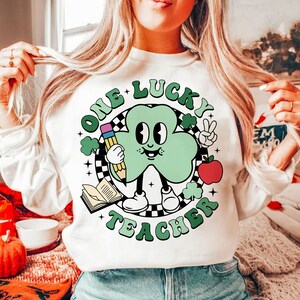 Pu&ograve; includere: Felpa bianca con un personaggio di cartone animato a forma di trifoglio verde che tiene una matita e una mela. Il testo "One Lucky Teacher" &egrave; scritto in un cerchio attorno al personaggio.