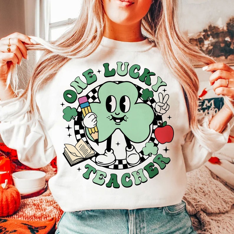 Pu&ograve; includere: Felpa bianca con un personaggio di cartone animato a forma di trifoglio verde che tiene una matita e una mela. Il testo "One Lucky Teacher" &egrave; scritto in un cerchio attorno al personaggio.