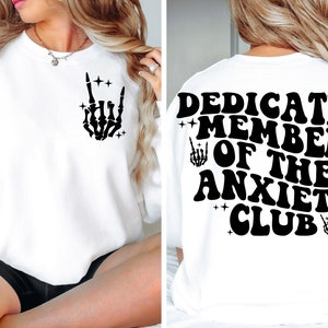 May include: White sweatshirt with black text and graphic. The front of the sweatshirt has a black skeleton hand making a rock and roll sign. The back of the sweatshirt says "Dedicated Member of the Anxiety Club" with a skeleton hand graphic on each side of the text.