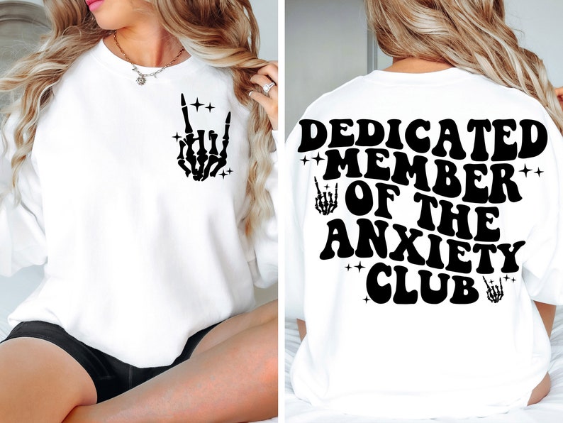 May include: White sweatshirt with black text and graphic. The front of the sweatshirt has a black skeleton hand making a rock and roll sign. The back of the sweatshirt says "Dedicated Member of the Anxiety Club" with a skeleton hand graphic on each side of the text.