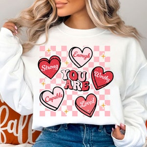 May include: White sweatshirt with a pink and white checkered background. The sweatshirt has red and pink hearts with the words "You Are" in pink and white letters. The hearts have the words "Strong", "Capable", "Loved", "Worthy", and "Enough" on them.