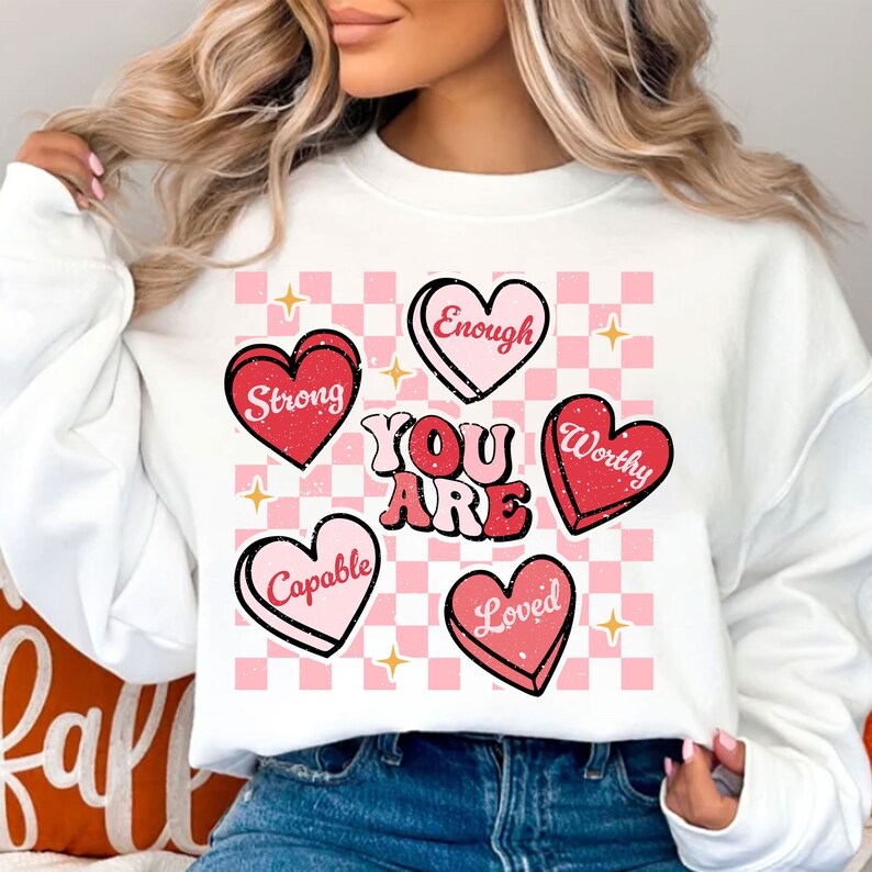 May include: White sweatshirt with a pink and white checkered background. The sweatshirt has red and pink hearts with the words "You Are" in pink and white letters. The hearts have the words "Strong", "Capable", "Loved", "Worthy", and "Enough" on them.