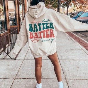 May include: A cream-colored hoodie with a retro baseball-themed graphic on the back. The graphic says "Hey Batter Batter Swing" with a baseball and stars.
