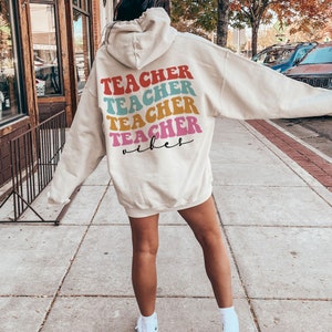 以下が含まれることがあります： クリーム色のフード付きスウェットシャツに、「TEACHER」という単語が虹色のカラーで4回繰り返されています。テキストはレトロなフォントで、「TEACHER」の単語の下に「vibes」という単語が黒で書かれています。