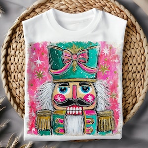 K&ouml;nnte beinhalten: Wei&szlig;es T-Shirt mit einer farbenfrohen Illustration eines Nussknacker-Soldaten. Das Design zeigt einen t&uuml;rkisfarbenen Hut mit einer rosa Schleife, goldfarbenen Akzenten und einem rosa-wei&szlig;en Hintergrund mit Sternen. Das T-Shirt wird auf einem geflochtenen Korb pr&auml;sentiert.