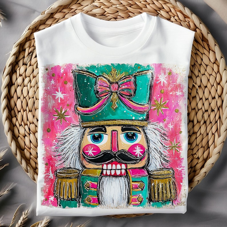 K&ouml;nnte beinhalten: Wei&szlig;es T-Shirt mit einer farbenfrohen Illustration eines Nussknacker-Soldaten. Das Design zeigt einen t&uuml;rkisfarbenen Hut mit einer rosa Schleife, goldfarbenen Akzenten und einem rosa-wei&szlig;en Hintergrund mit Sternen. Das T-Shirt wird auf einem geflochtenen Korb pr&auml;sentiert.