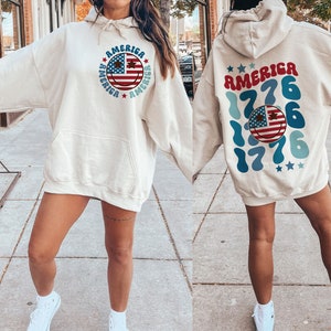 Pode incluir: Uma camisola de capuz cor de creme com um design de cara sorridente que apresenta a bandeira americana e o texto "America" repetido em torno do rosto. A parte de tr&aacute;s da camisola tem o texto "America 1776" repetido num design azul e branco.