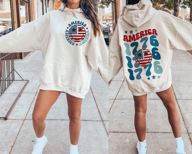 Pode incluir: Uma camisola de capuz cor de creme com um design de cara sorridente que apresenta a bandeira americana e o texto "America" repetido em torno do rosto. A parte de tr&aacute;s da camisola tem o texto "America 1776" repetido num design azul e branco.