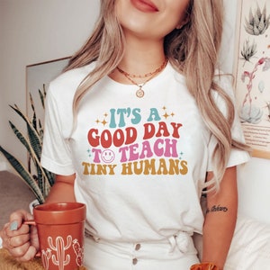以下が含まれることがあります： 「It's a good day to teach tiny humans」とレトロなスタイルのグラフィックがプリントされた白いTシャツ。グラフィックは虹色で、笑顔の顔があります。