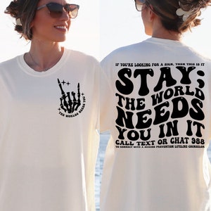 Puede incluir: Camiseta color crema con un gr&aacute;fico de mano de esqueleto negro haciendo un gesto de rock and roll, y el texto "THE WORLD NEEDS YOU". La parte trasera de la camiseta presenta el texto "STAY; THE WORLD NEEDS YOU IN IT" y un n&uacute;mero de tel&eacute;fono.
