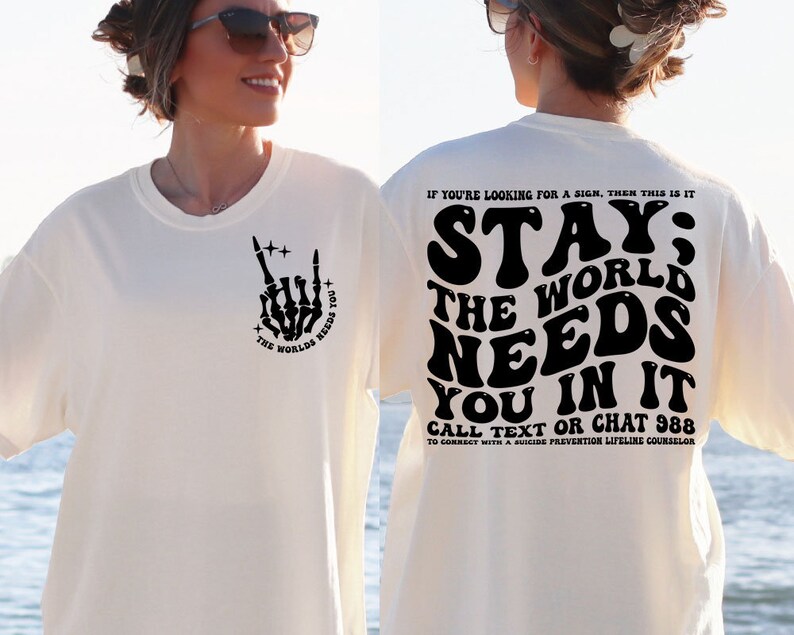 Puede incluir: Camiseta color crema con un gr&aacute;fico de mano de esqueleto negro haciendo un gesto de rock and roll, y el texto "THE WORLD NEEDS YOU". La parte trasera de la camiseta presenta el texto "STAY; THE WORLD NEEDS YOU IN IT" y un n&uacute;mero de tel&eacute;fono.