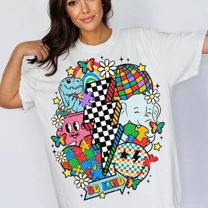 Op de afbeelding: Wit T-shirt met een kleurrijk grafisch ontwerp met een bliksem, een dinosaurus, een puzzelhart, een smiley, een madeliefje, een vlinder, een regenboog, een discobal en een olifant. De tekst "BE KIND" is op de grafiek gedrukt.