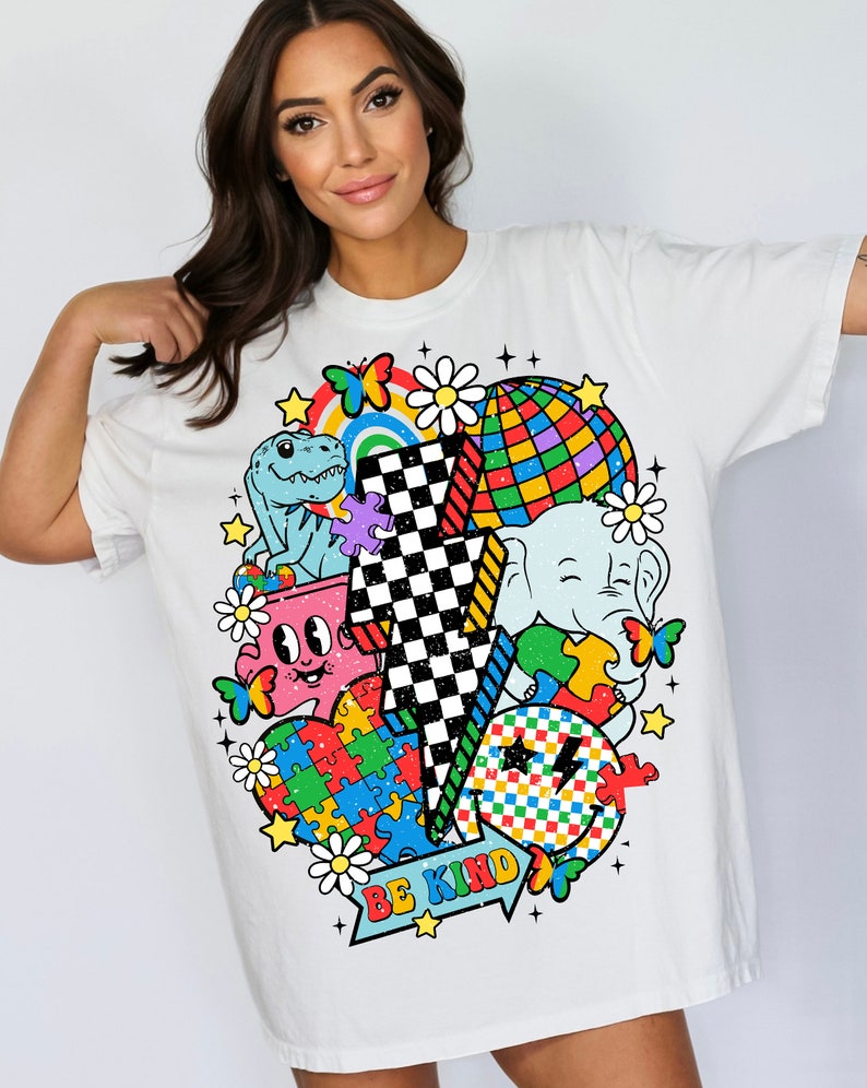 Op de afbeelding: Wit T-shirt met een kleurrijk grafisch ontwerp met een bliksem, een dinosaurus, een puzzelhart, een smiley, een madeliefje, een vlinder, een regenboog, een discobal en een olifant. De tekst "BE KIND" is op de grafiek gedrukt.