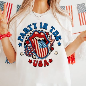 May include: White t-shirt with a red, white, and blue graphic design. The graphic features a tongue with the American flag design, a disco ball, and the text "Party in the USA".