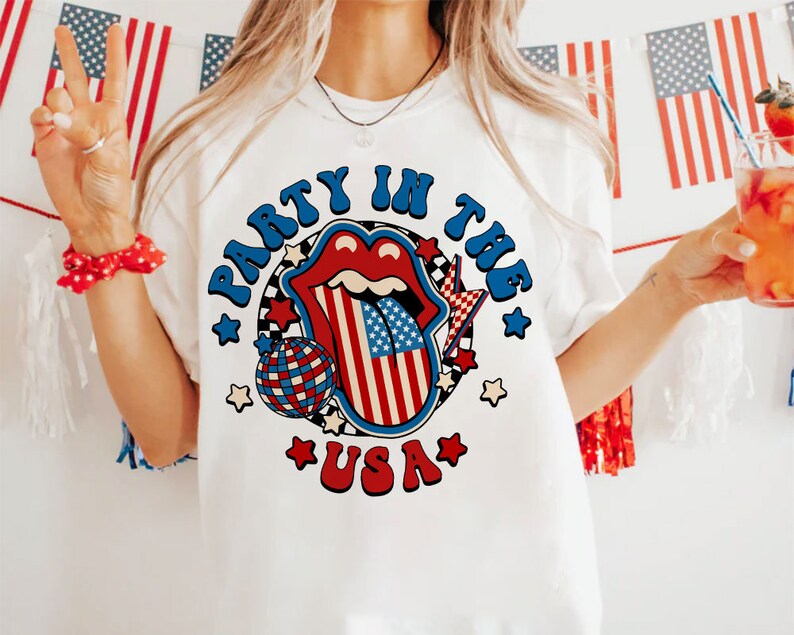 May include: White t-shirt with a red, white, and blue graphic design. The graphic features a tongue with the American flag design, a disco ball, and the text "Party in the USA".