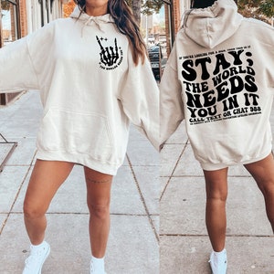 Puede incluir: Sudadera con capucha color crema con bolsillo delantero y capucha. La parte delantera presenta un gr&aacute;fico de mano de esqueleto negro con el texto "THE WORLD NEEDS YOU". La parte trasera muestra el texto "STAY; THE WORLD NEEDS YOU IN IT".