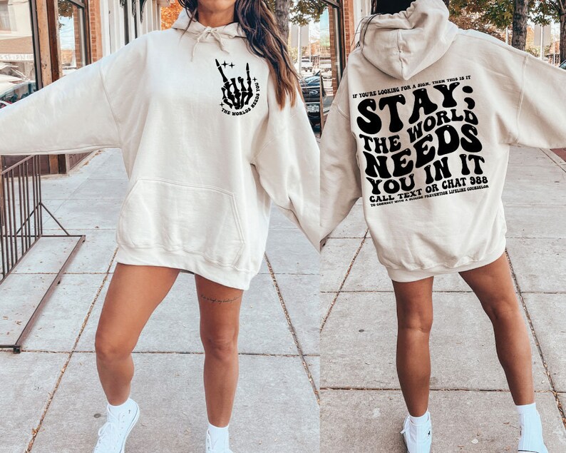 Puede incluir: Sudadera con capucha color crema con bolsillo delantero y capucha. La parte delantera presenta un gr&aacute;fico de mano de esqueleto negro con el texto "THE WORLD NEEDS YOU". La parte trasera muestra el texto "STAY; THE WORLD NEEDS YOU IN IT".