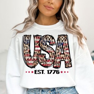 May include: White sweatshirt with a graphic of the word "USA" in red, white, and blue with a leopard print pattern. The graphic also includes the text "EST. 1776" with stars.