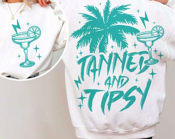 Op de afbeelding: Witte trui met een turquoise palmboom, bliksemschichten en twee margarita glazen. De tekst "TANNED AND TIPSY" is in turquoise geschreven.