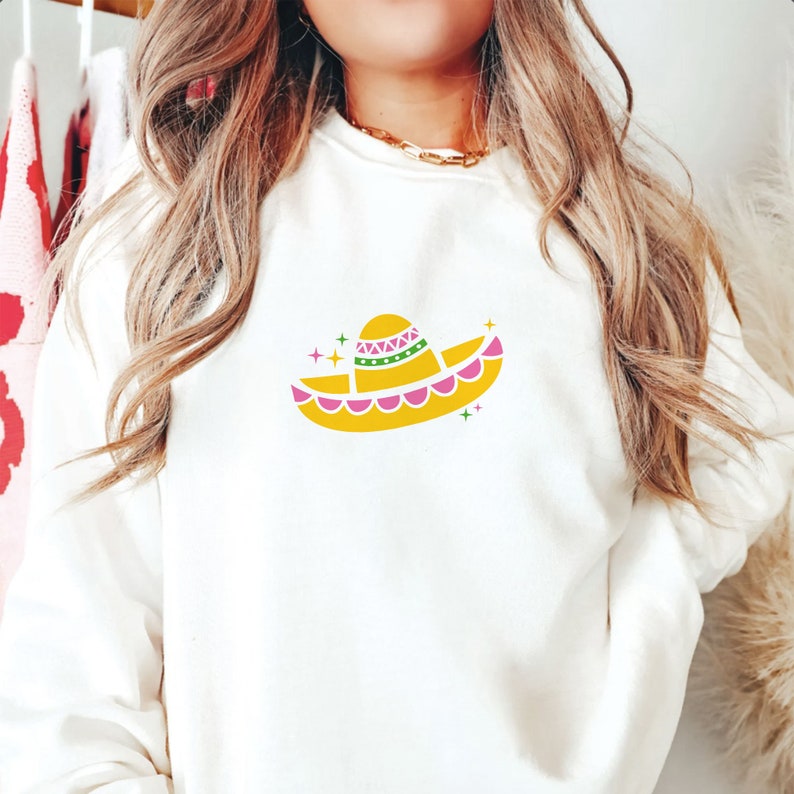 May include: White sweatshirt with a yellow sombrero graphic. The sombrero has pink and green accents and is surrounded by small white stars.