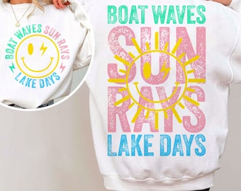 Op de afbeelding: Witte sweatshirt met een grafisch ontwerp met een gele smiley met bliksemschichten en de woorden "Boat Waves, Sun Rays, Lake Days". De achterkant van de sweatshirt heeft hetzelfde ontwerp met het woord "Sundays".