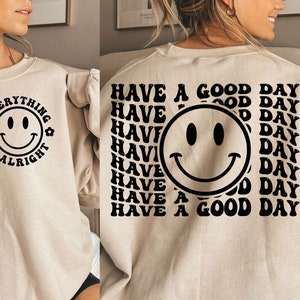 K&ouml;nnte beinhalten: Ein beiger Pullover mit einem schwarzen Smiley und dem Text "Everything is alright" auf der Vorderseite. Die R&uuml;ckseite des Pullovers hat einen schwarzen Smiley und den Text "Have a good day" mehrmals wiederholt.