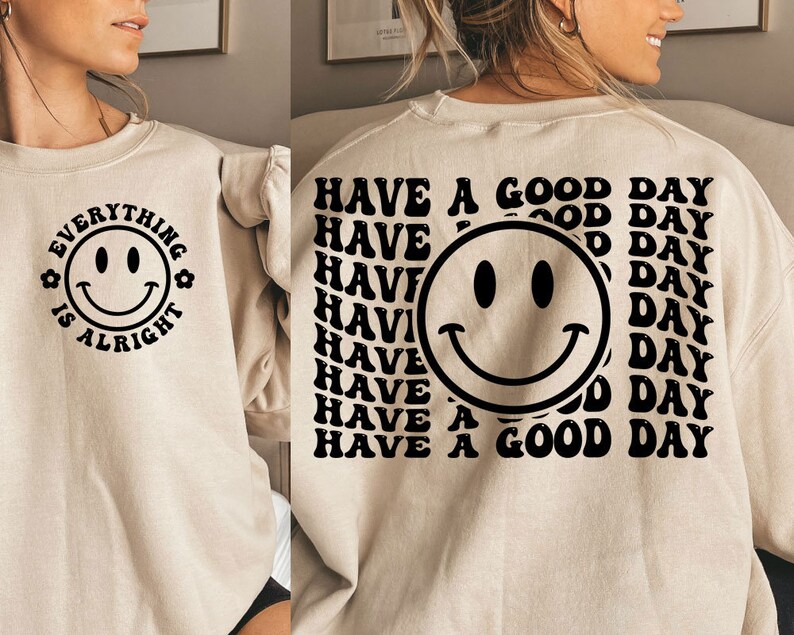 K&ouml;nnte beinhalten: Ein beiger Pullover mit einem schwarzen Smiley und dem Text "Everything is alright" auf der Vorderseite. Die R&uuml;ckseite des Pullovers hat einen schwarzen Smiley und den Text "Have a good day" mehrmals wiederholt.