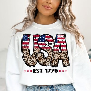 May include: White sweatshirt with a graphic of the word "USA" in large, bold letters. The letters are decorated with an American flag design and a leopard print pattern. The text "EST. 1776" is printed below the graphic.