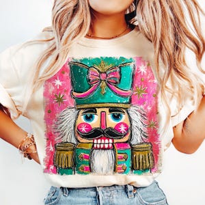 K&ouml;nnte beinhalten: Cremefarbenes Sweatshirt mit einer farbenfrohen, skurrilen Illustration eines Nussknackers. Der Nussknacker hat einen gr&uuml;nen Hut mit einer rosa Schleife, einen schwarzen Schnurrbart und blaue Augen. Der Hintergrund ist rosa mit goldenen Sternen.