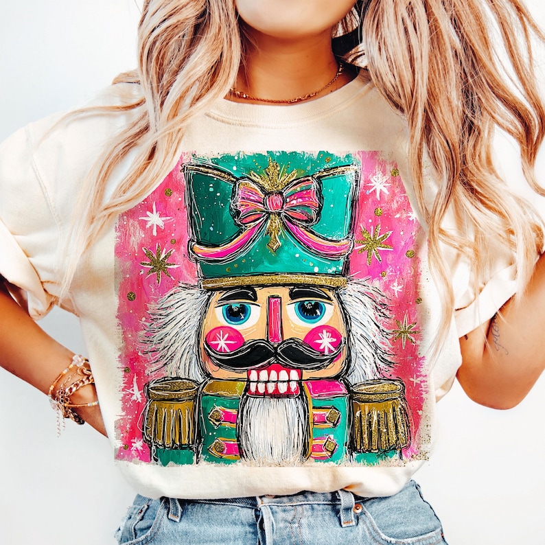 K&ouml;nnte beinhalten: Cremefarbenes Sweatshirt mit einer farbenfrohen, skurrilen Illustration eines Nussknackers. Der Nussknacker hat einen gr&uuml;nen Hut mit einer rosa Schleife, einen schwarzen Schnurrbart und blaue Augen. Der Hintergrund ist rosa mit goldenen Sternen.